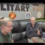 Two people sit on a black couch in an interview setting A woman with glasses and a ponytail listens attentively A bald man gestures as he speaks Military emblems are displayed on the wall behind them A blanket with "Good Vibes" is draped over the couch